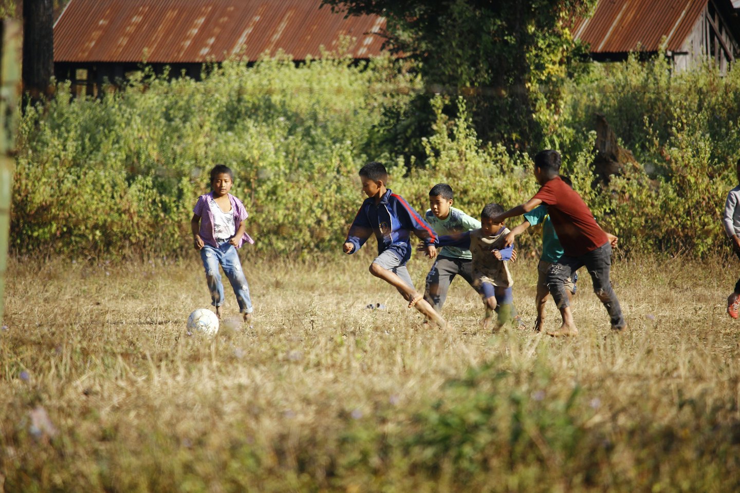 Children playing soccer on a grassy field with rustic buildings.