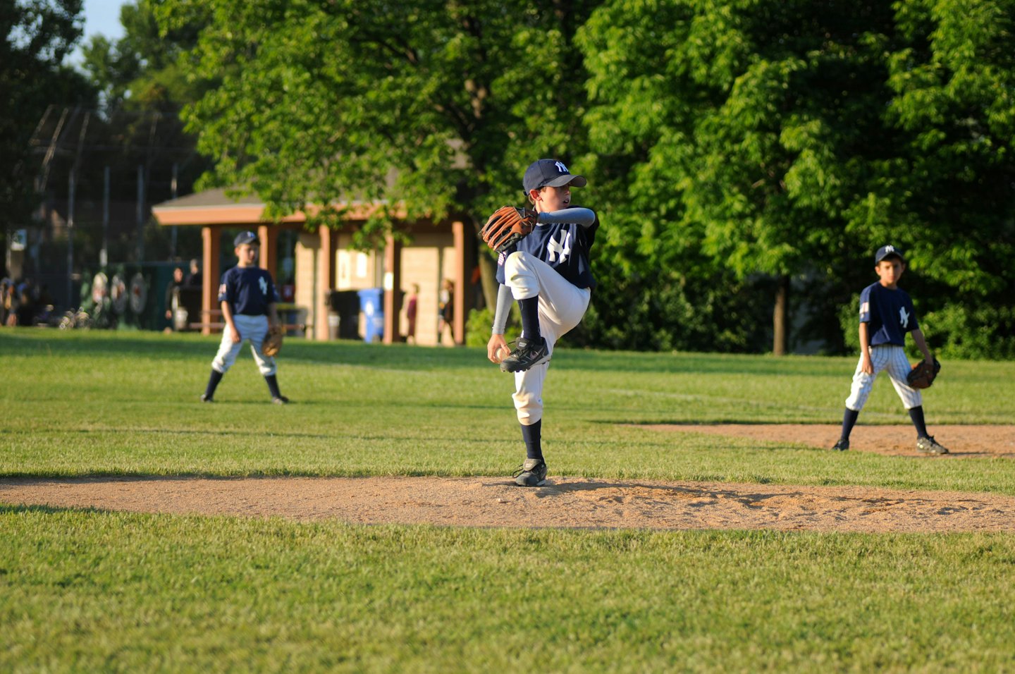 Three boys in baseball uniforms pitching and fielding on a sunny field.
